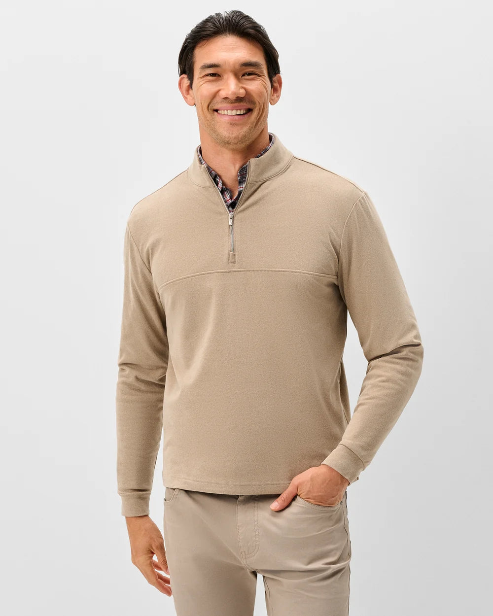 A smiling man wearing a beige long-sleeve quarter-zip pullover sweater over a patterned collared shirt, paired with matching beige pants, against a light gray studio background.