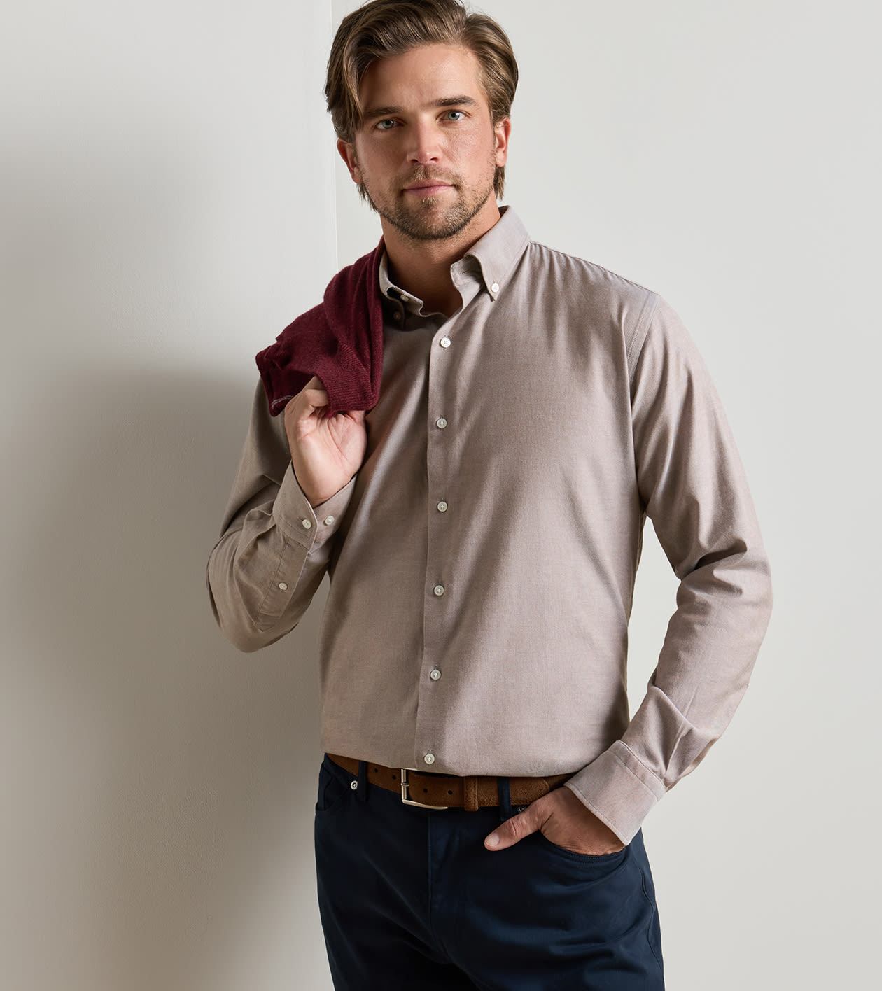 A man wearing a light taupe long-sleeve button-down dress shirt, dark navy trousers, and a brown belt, holding a dark burgundy shirt under his arm against a light gray studio background.