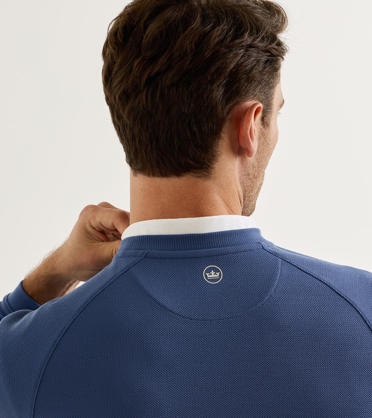 Rear view of a man wearing a textured blue pullover sweater with a white collar, showing a small embroidered crown logo on the back neck.