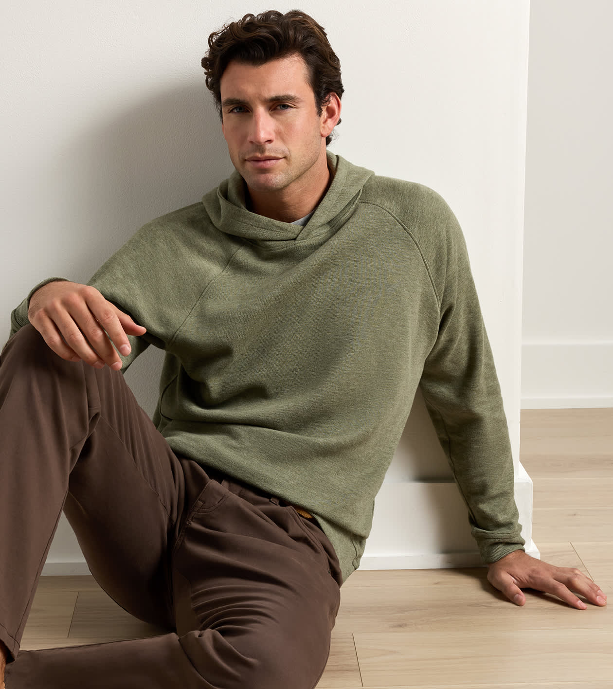 A man wearing an olive green raglan hoodie and brown pants sits leaning against a white wall on a light wood floor.