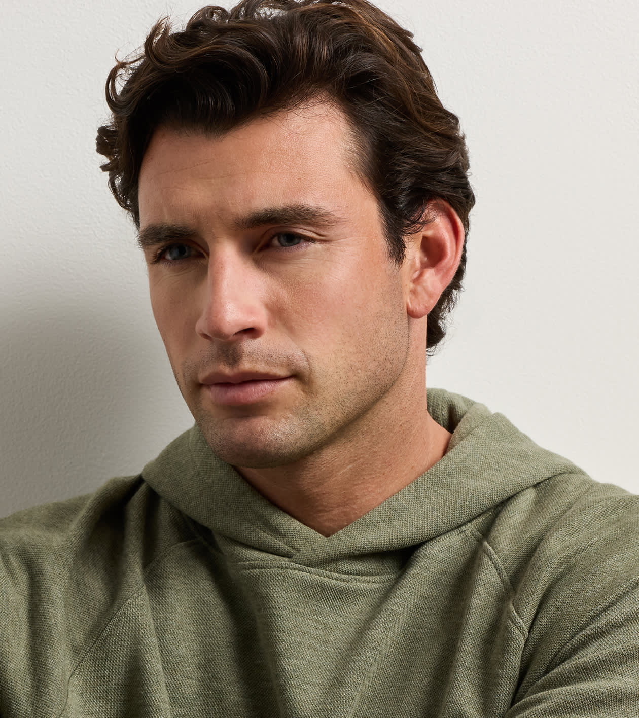 A close-up portrait of a man with wavy brown hair wearing an olive green hooded sweatshirt, set against a plain light gray background.