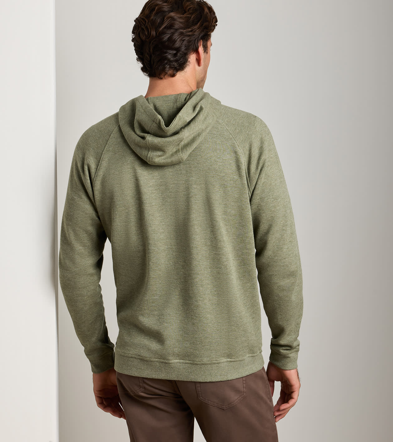 A man viewed from behind wearing an olive green hooded sweatshirt and brown trousers, standing against a plain light gray wall.