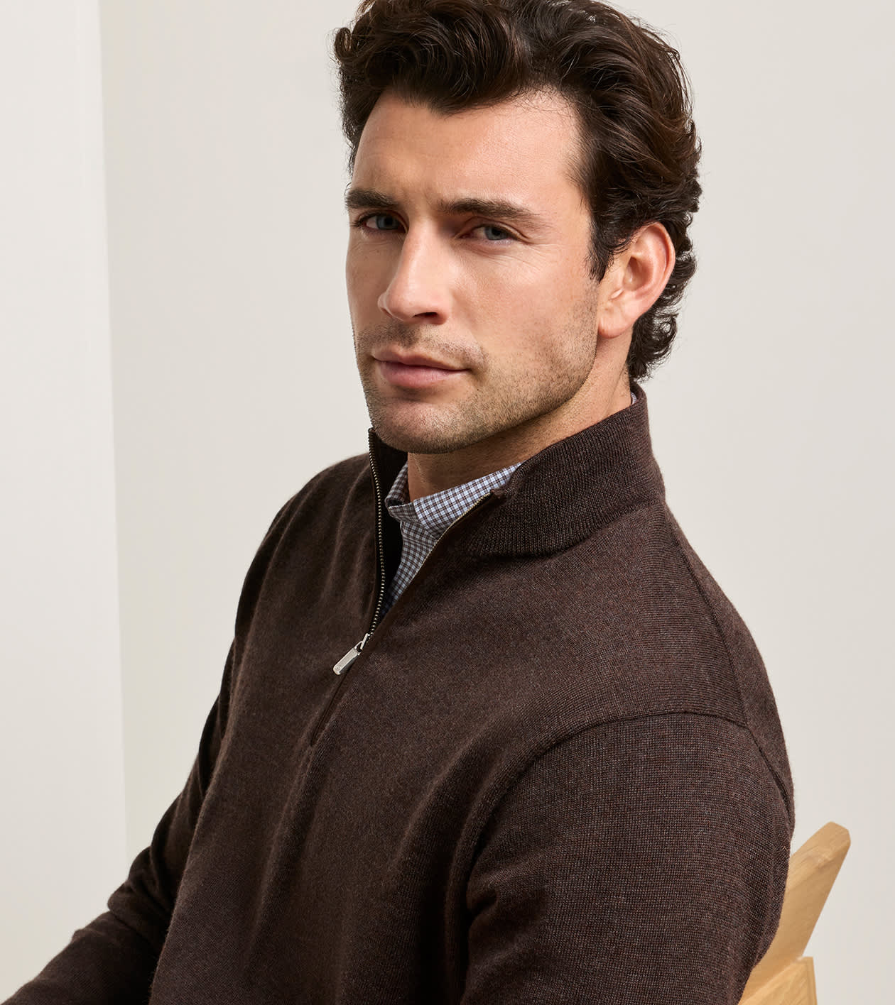 A man wearing a dark brown quarter-zip knit sweater over a small checkered shirt, seated against a light neutral background.