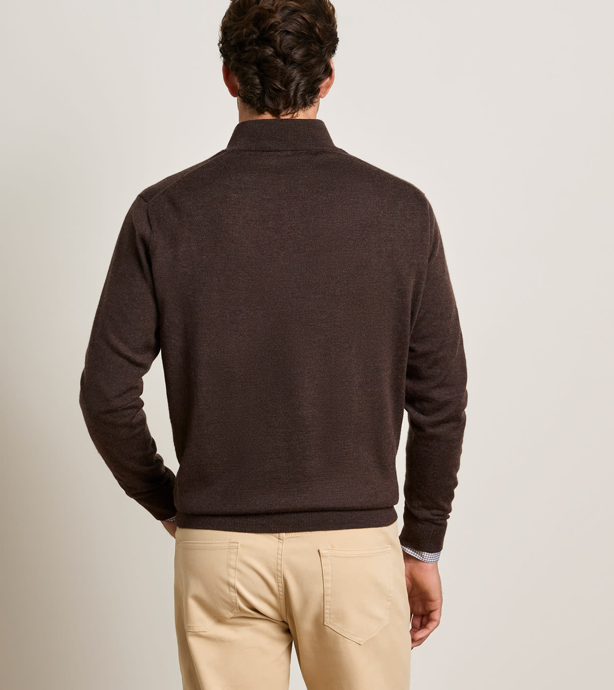 A man viewed from behind wearing a dark brown mock neck knit sweater and beige trousers, standing against a plain light gray studio background.