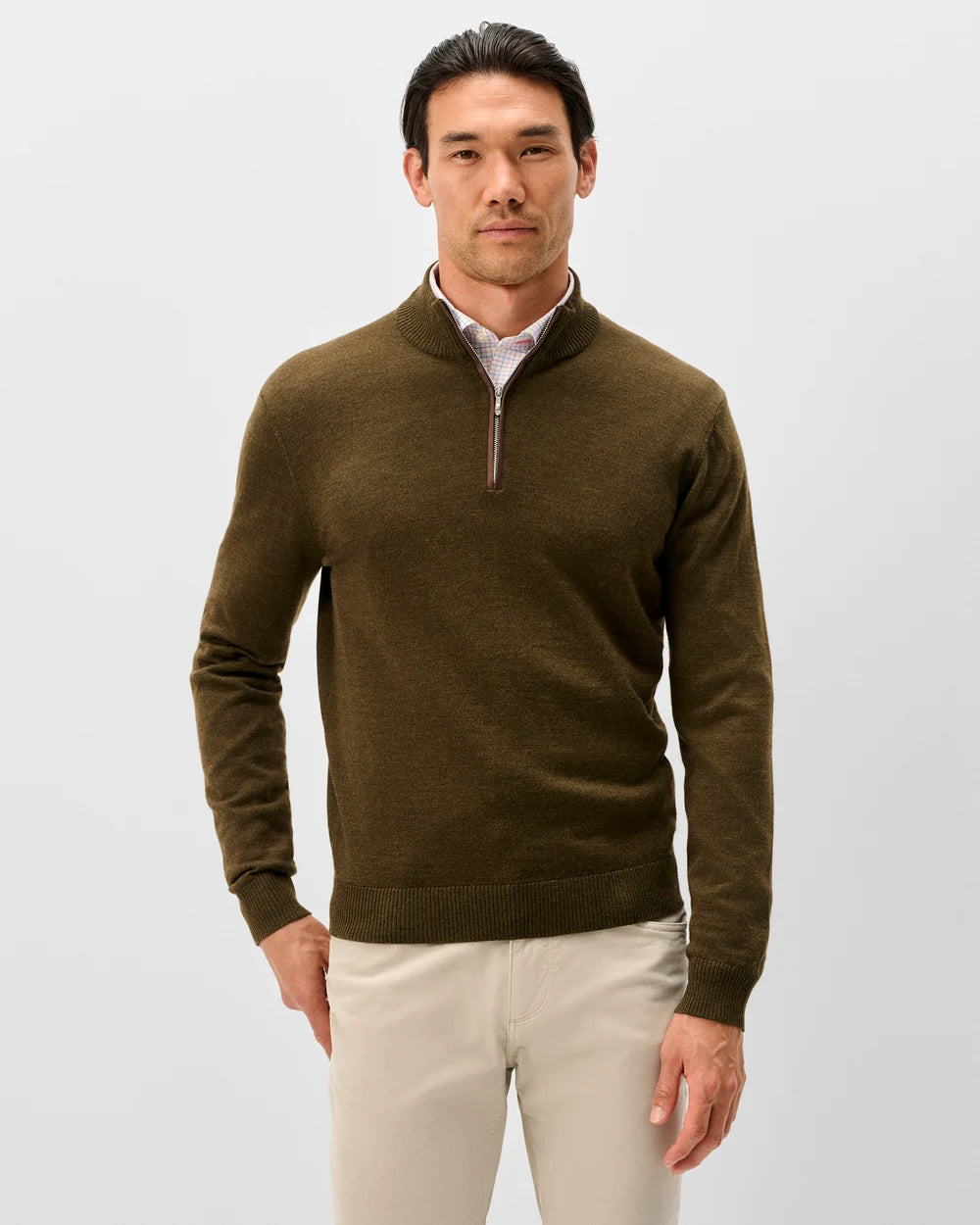 A man wearing a dark olive brown quarter-zip knit sweater over a light pink collared shirt and beige trousers, standing against a light gray studio background.