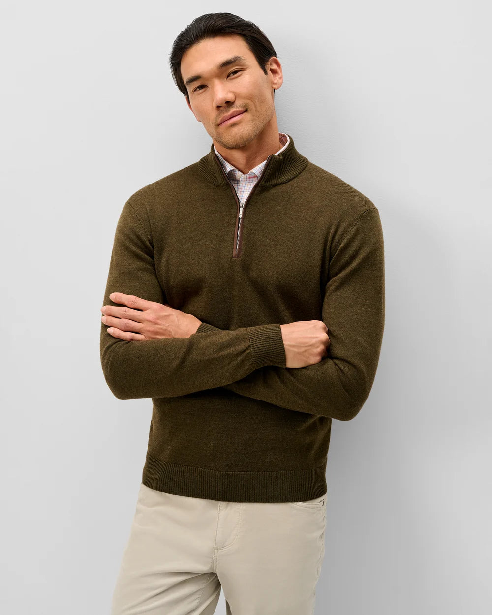 A man wearing an olive green quarter-zip knit sweater over a light pink collared shirt and beige trousers, standing against a light gray studio background.