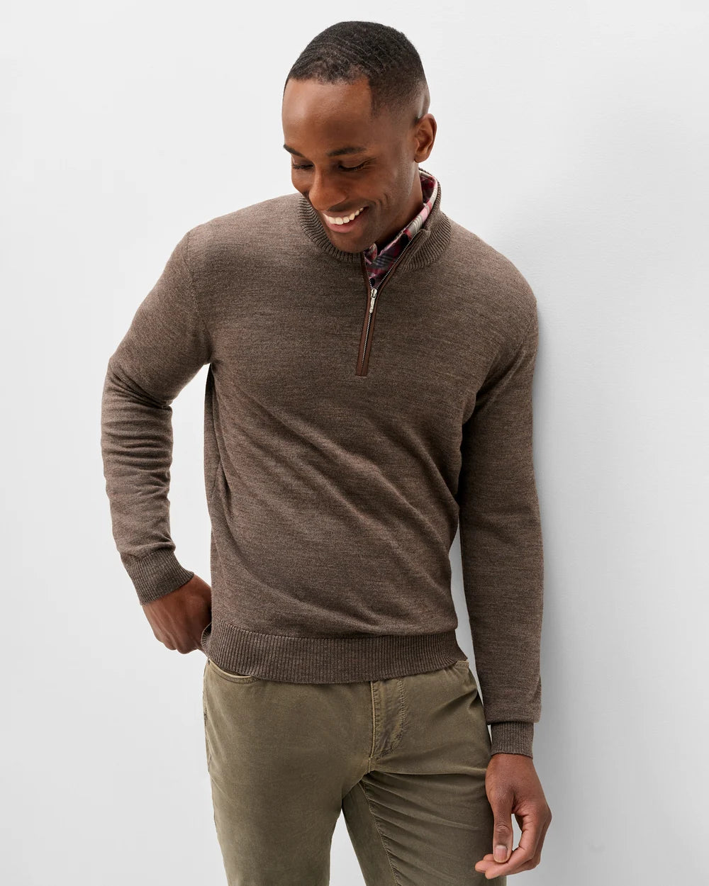 A man wearing a brown quarter-zip knit sweater over a patterned collared shirt and olive green trousers, standing against a light gray studio background.