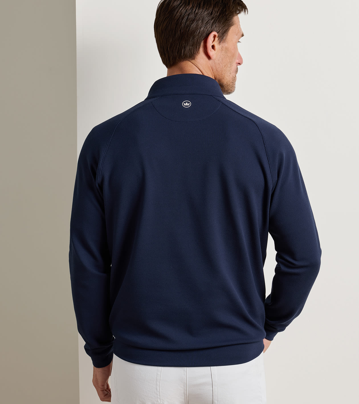A man shown from the back wearing a navy blue quarter-zip pullover sweater and white pants, standing against a neutral light gray wall.