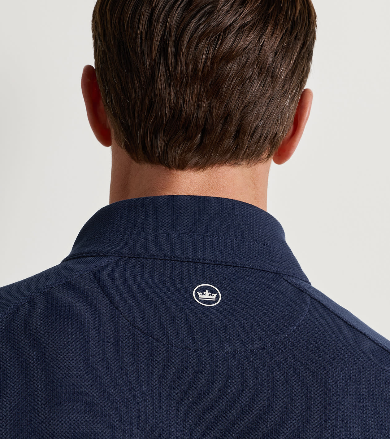 Close-up back view of a man wearing a navy blue textured polo shirt, showing the collar and a small white embroidered crown logo.