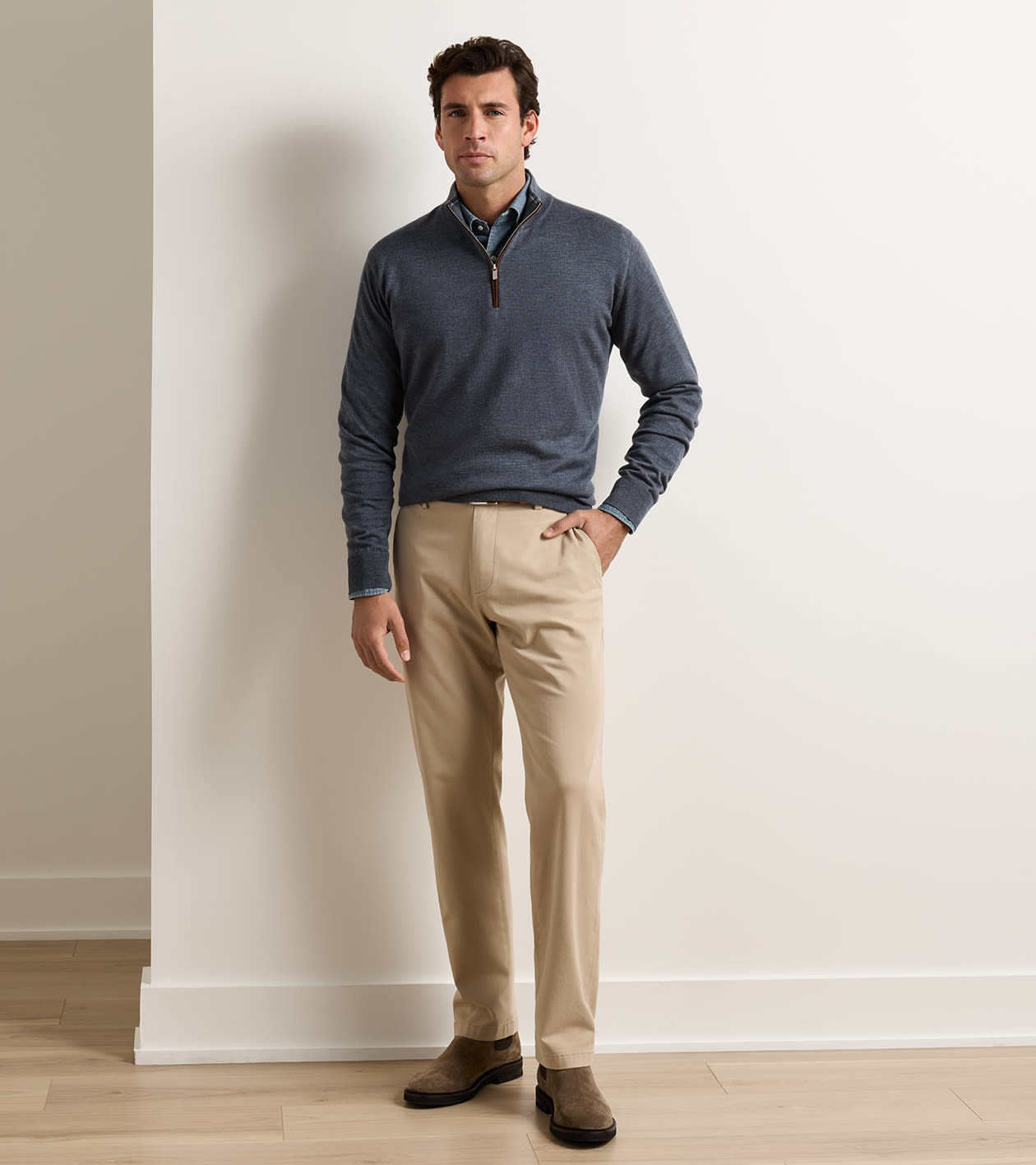 A man modeling a slate blue quarter-zip sweater over a collared shirt and beige trousers, standing against a white wall on a light wood floor.