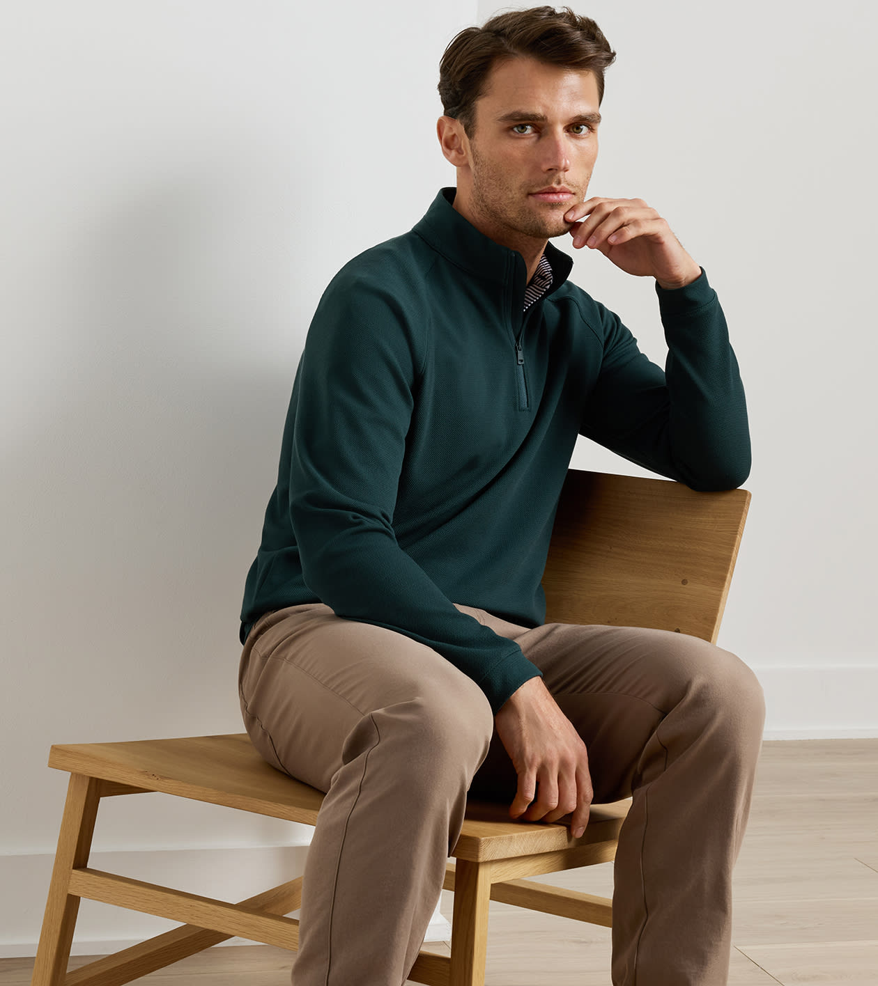 A man wearing a dark teal green quarter-zip long-sleeve shirt and tan pants, sitting on a light wood chair against a neutral off-white wall.