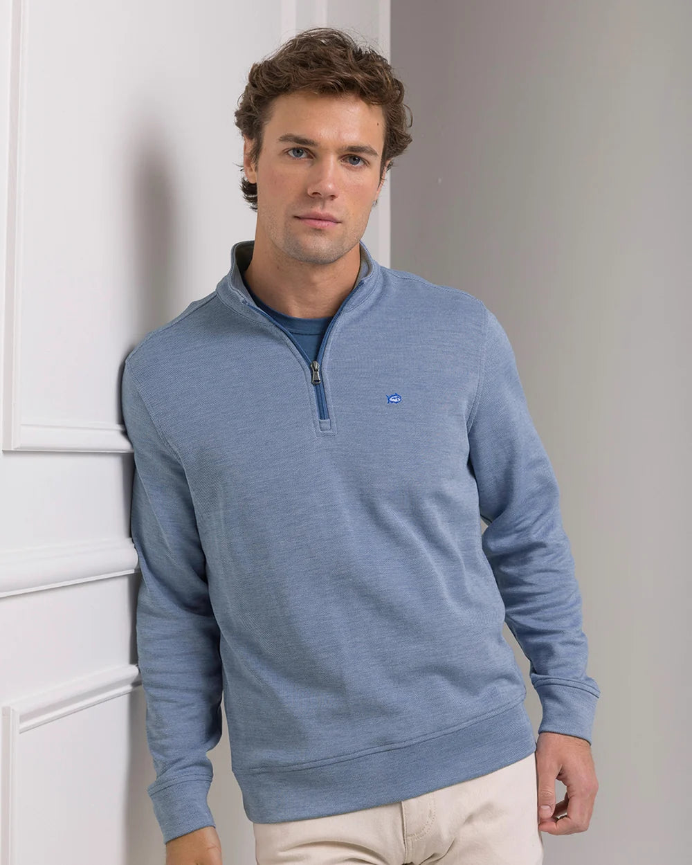 A man wearing a light blue quarter-zip long-sleeve sweater with a small blue whale logo on the chest, paired with beige pants, standing against a white paneled wall.