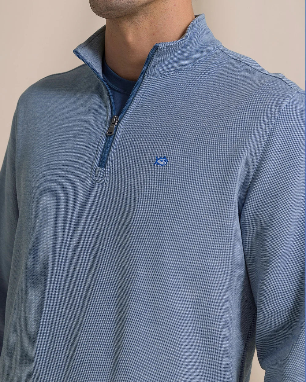 A close-up of a man wearing a heathered blue quarter-zip pullover sweater with a small blue embroidered logo on the chest, layered over a darker blue t-shirt.