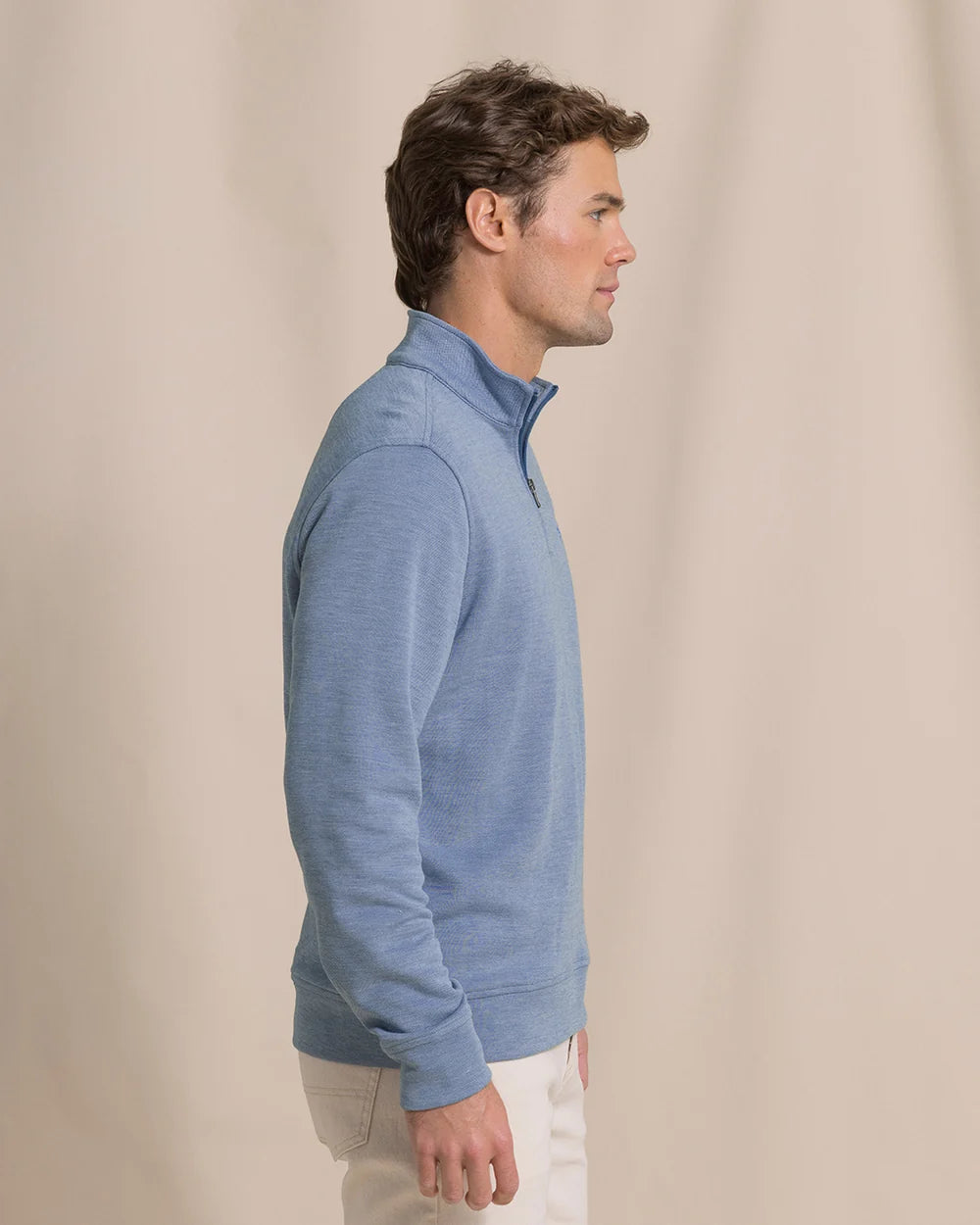 A man in profile wearing a light blue quarter-zip pullover sweater and off-white pants, standing against a plain beige studio backdrop.