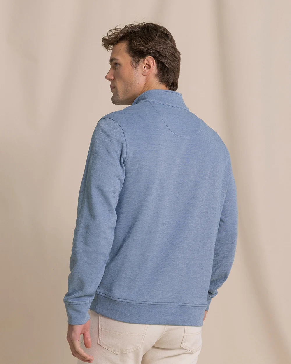 A man wearing a heathered light blue quarter-zip pullover sweatshirt, shown from a rear three-quarter angle against a neutral beige studio backdrop.