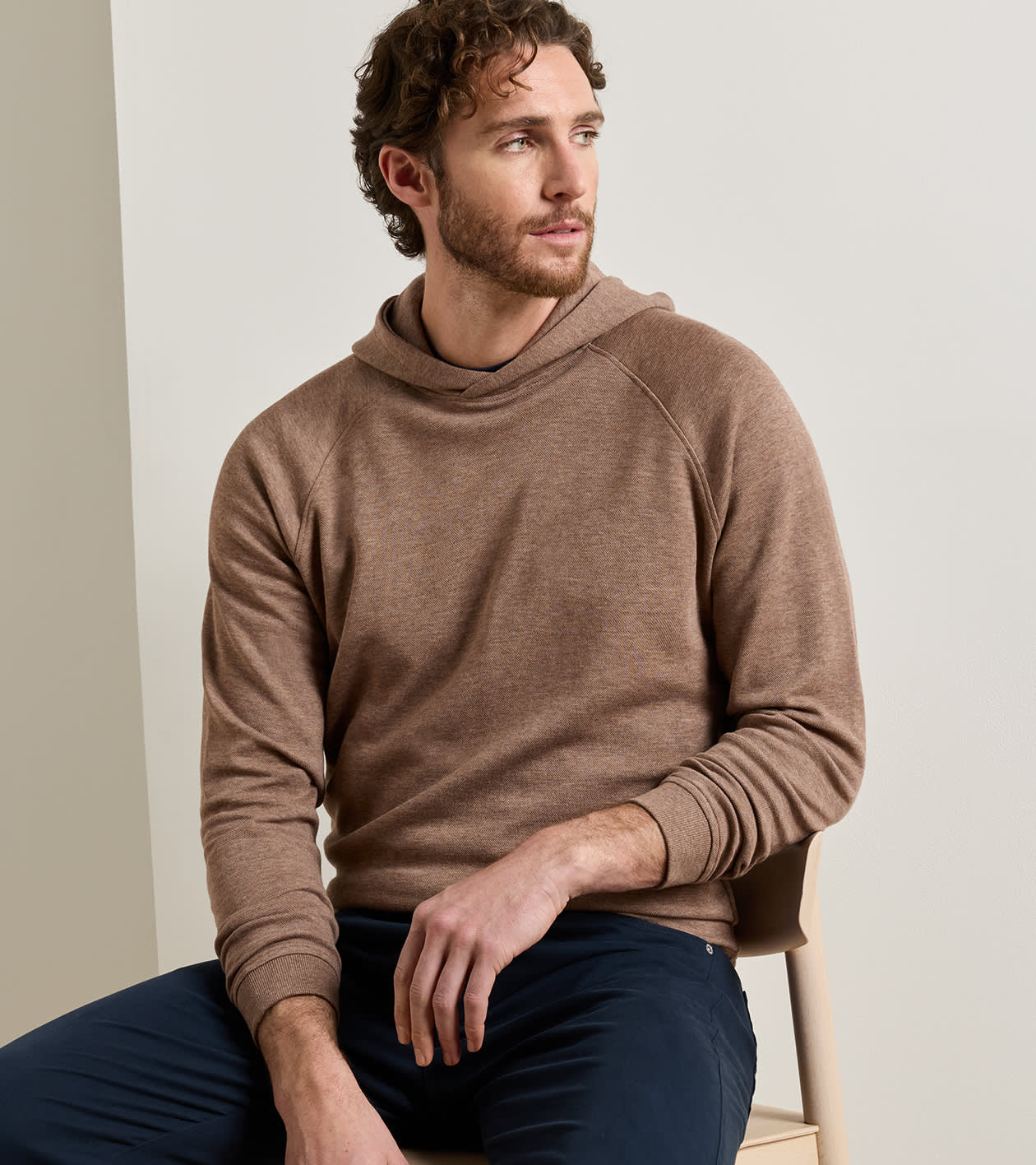 A man wearing a brown long-sleeve knit hoodie and navy pants, sitting on a light-colored wooden chair against a neutral beige background.