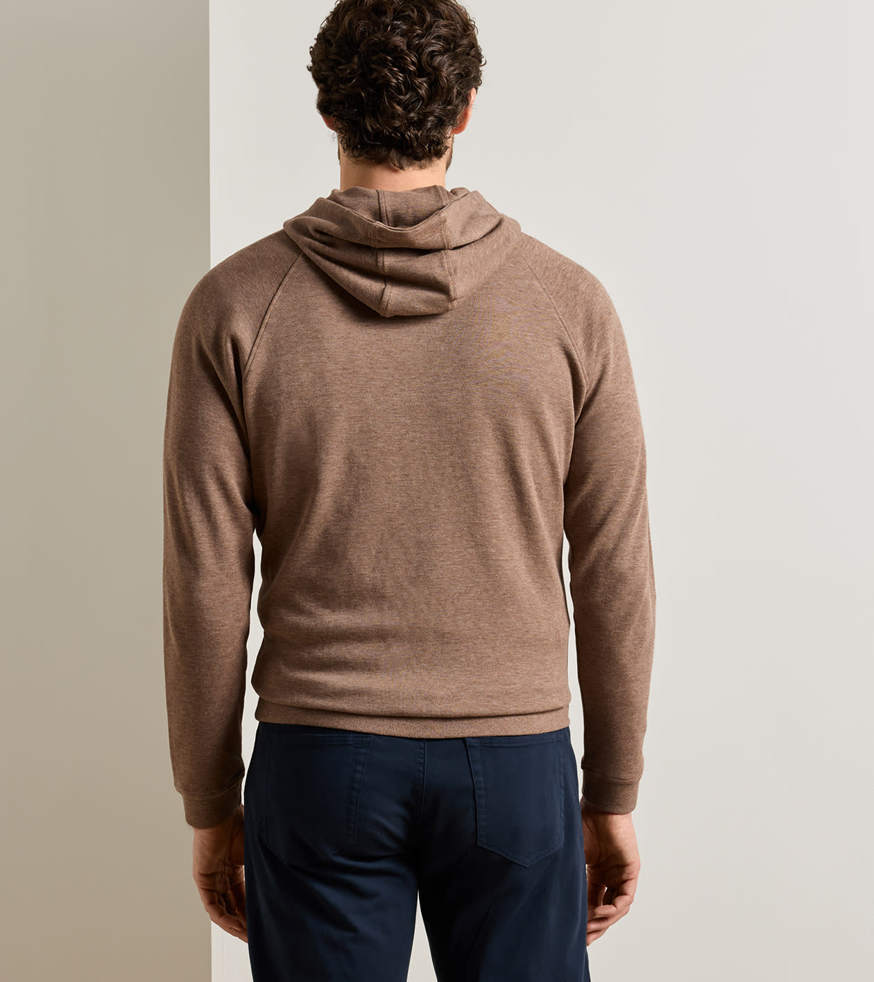 A man viewed from behind wearing a brown knit hooded sweater and navy blue trousers, standing against a neutral light gray background.
