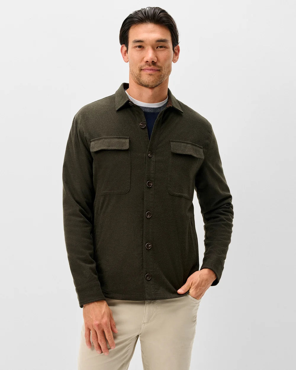 A man wearing a dark olive green long-sleeve button-up shirt with two chest flap pockets, layered over a navy blue t-shirt and paired with beige pants, standing against a light gray studio background.