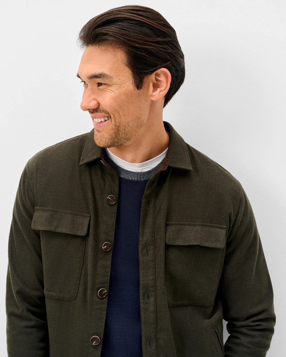 A man wearing an olive green button-up overshirt over a navy blue sweater and white undershirt, shown from the chest up against a light gray studio background.