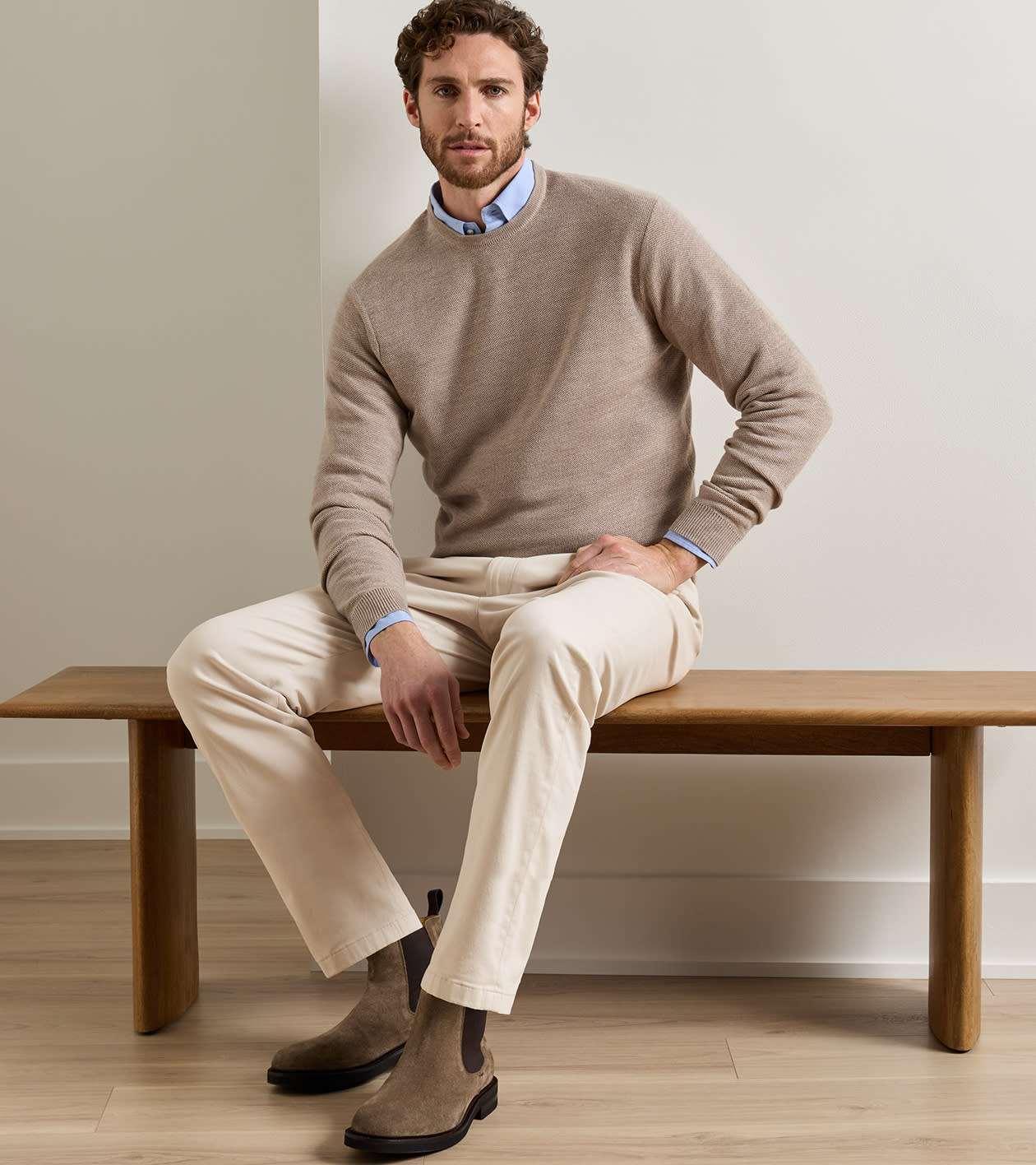 A man sits on a wooden bench wearing a taupe knit crewneck sweater over a light blue collared shirt, paired with beige trousers and brown Chelsea boots in a minimalist indoor setting.
