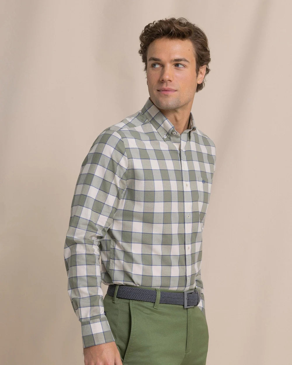 A man wearing a sage green and white checkered long-sleeve button-down shirt, paired with olive green trousers and a dark patterned belt, standing against a neutral beige background.