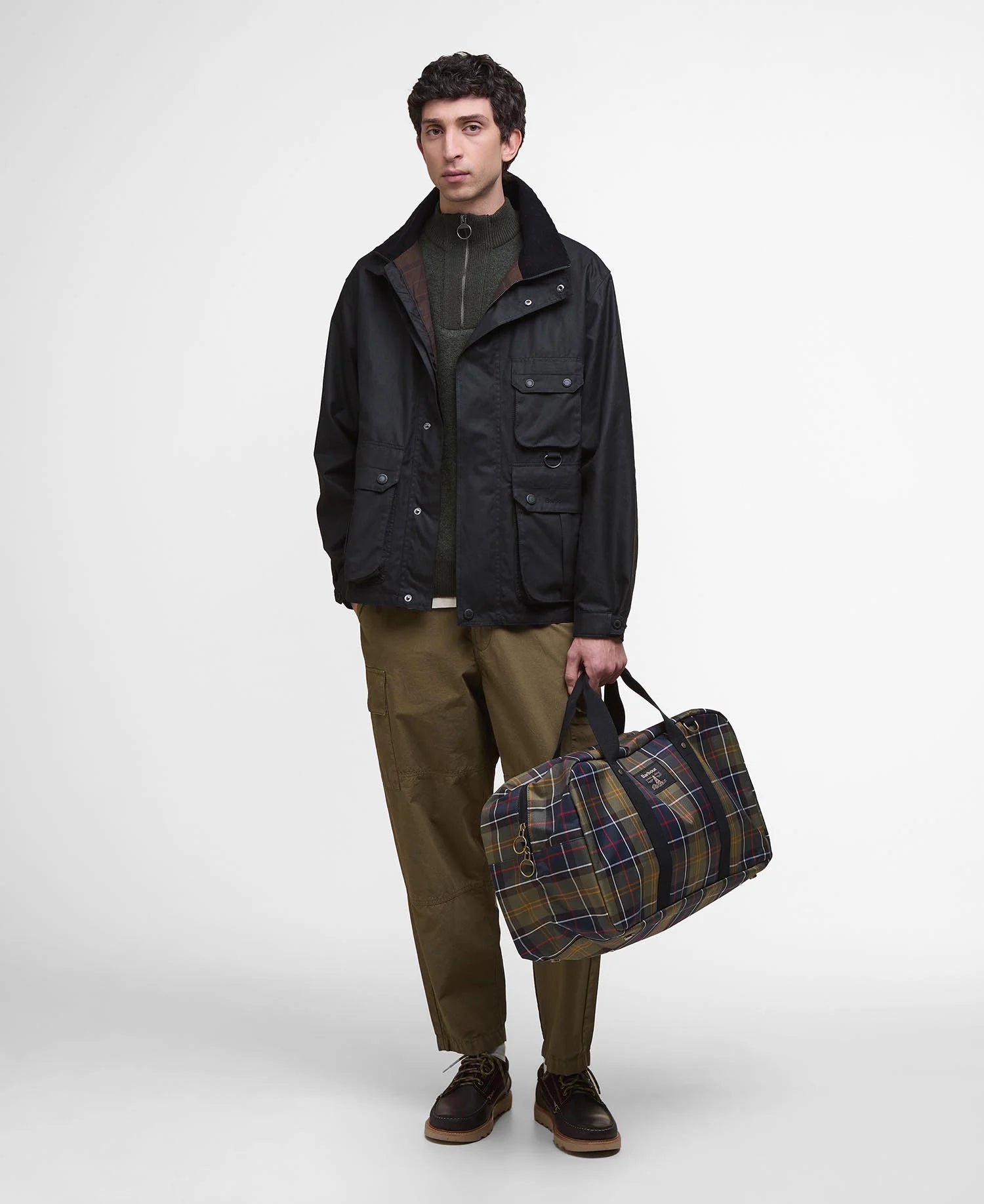 Man holding a plaid duffel bag wearing a dark jacket and brown pants on a white background