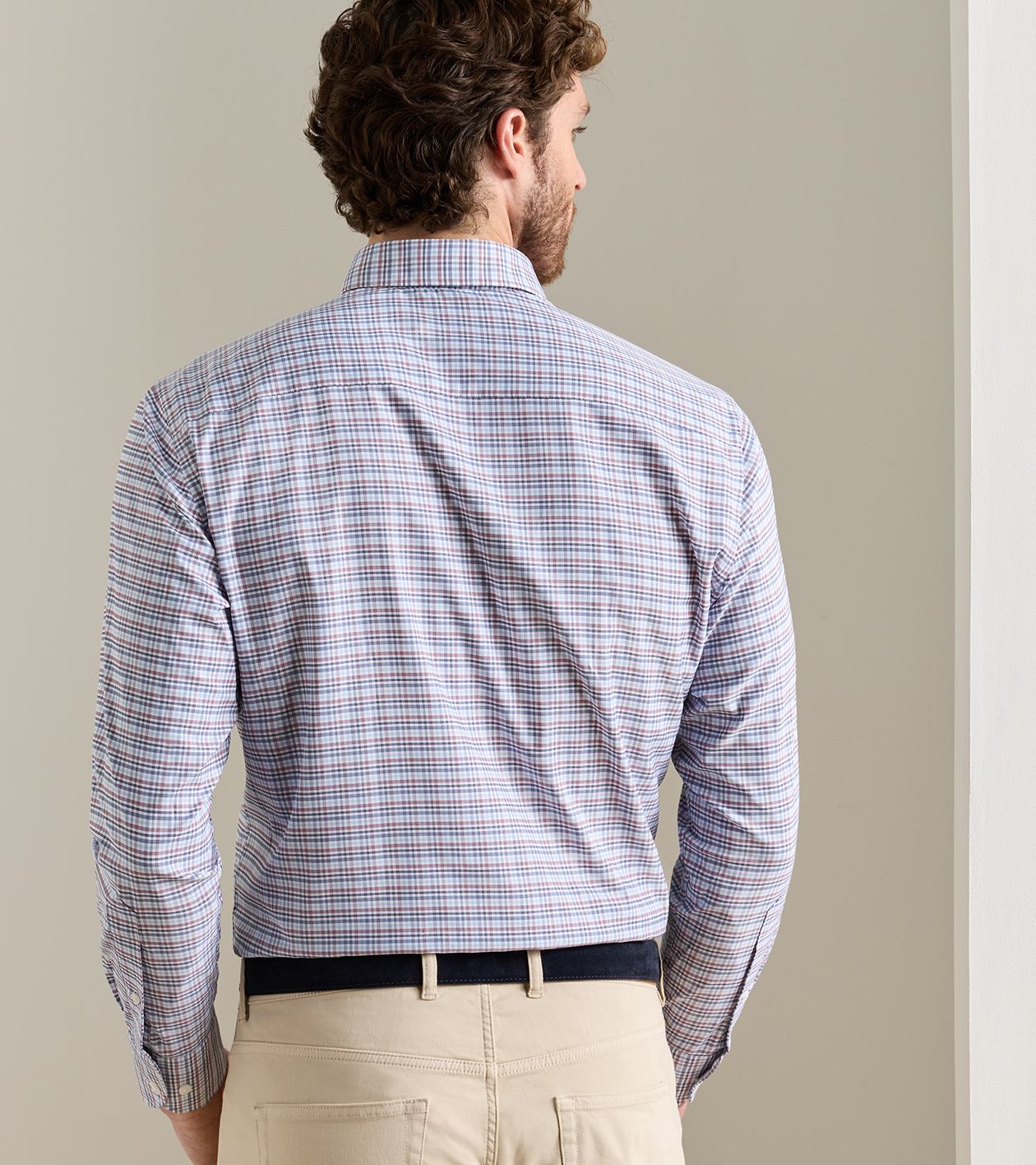 A man viewed from behind wearing a long-sleeve button-down shirt with a fine blue, red, and white plaid pattern, paired with khaki pants and a black belt.