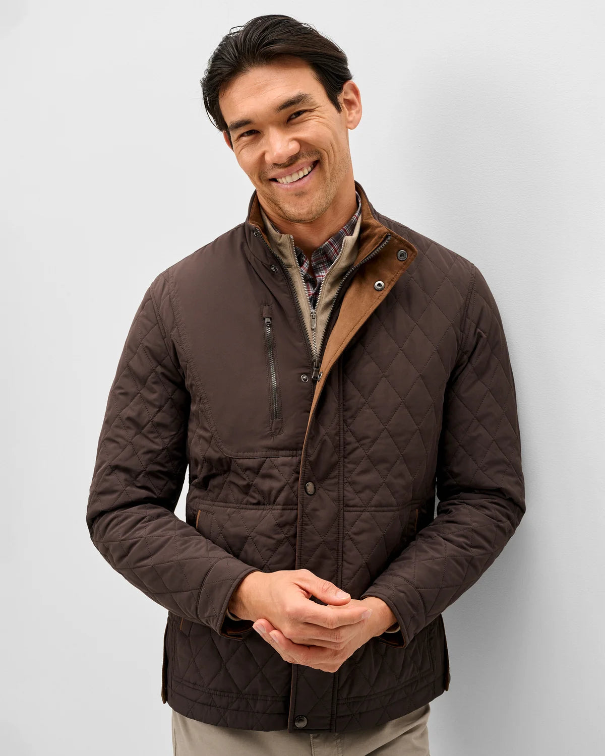 A smiling man wearing a dark brown diamond-quilted jacket with a tan collar lining, standing against a light gray studio background.