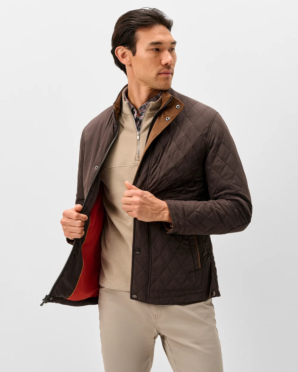 A man wearing a dark brown diamond-quilted jacket over a beige quarter-zip sweater and khaki pants, standing against a light gray studio background.