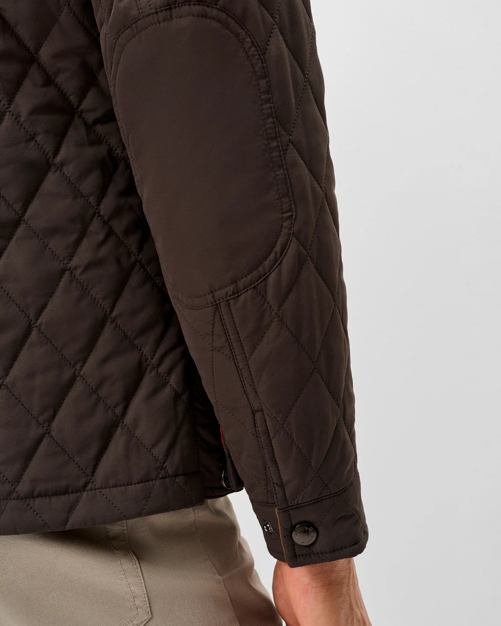 Close-up of a dark brown diamond-quilted jacket with elasticated elbow patches and a snap-button cuff, worn by a model with beige trousers against a light gray background.