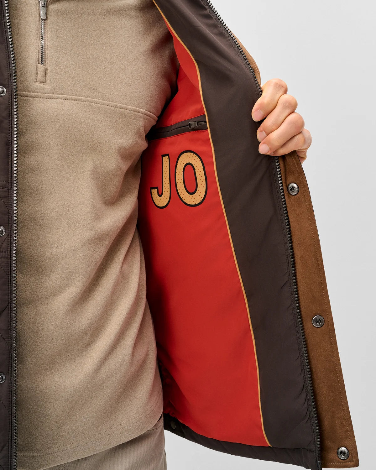 Close-up of a person wearing a tan jacket and opening a brown and red zippered layer to reveal a red inner pocket with gold 'JO' embroidery, on a light gray background.