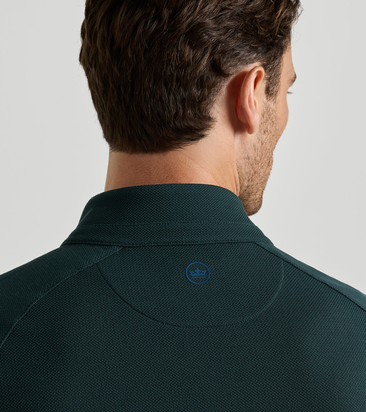 Close-up back view of a man wearing a dark green textured polo shirt, showing the collar and a small embroidered logo on the upper back.