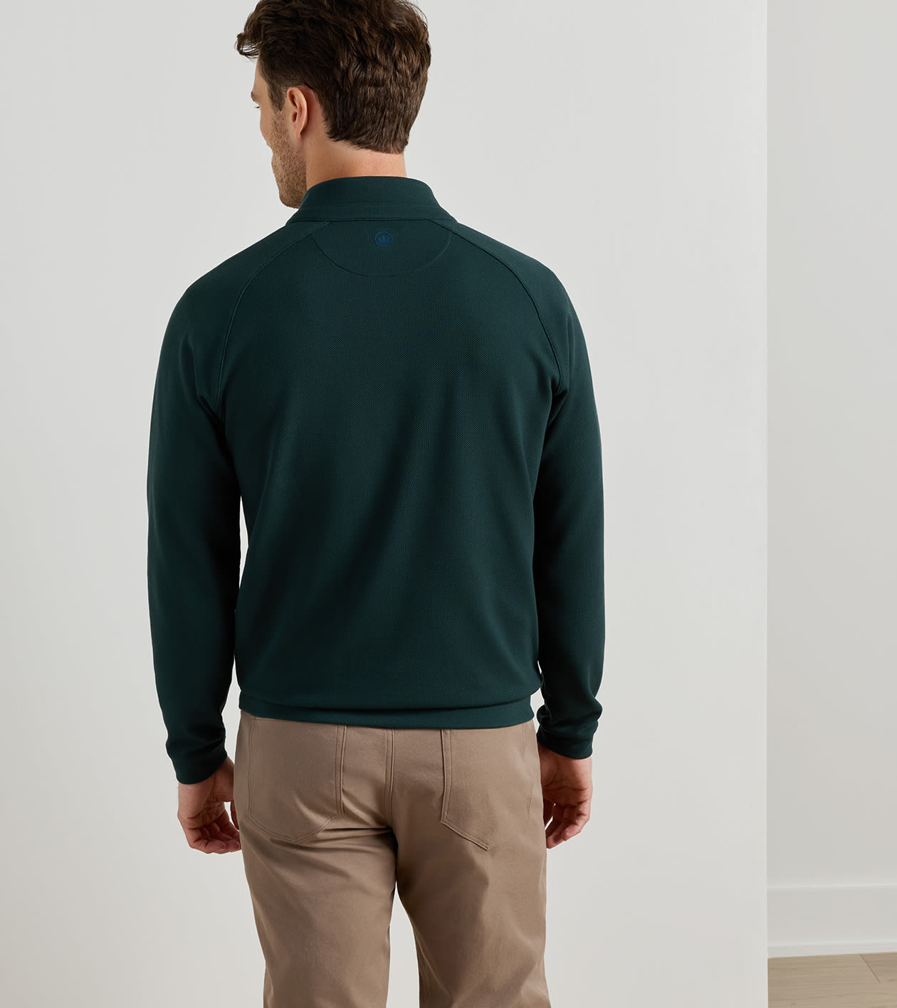 A man viewed from behind wearing a dark green long-sleeve quarter-zip pullover and tan pants, standing against a plain light gray wall.