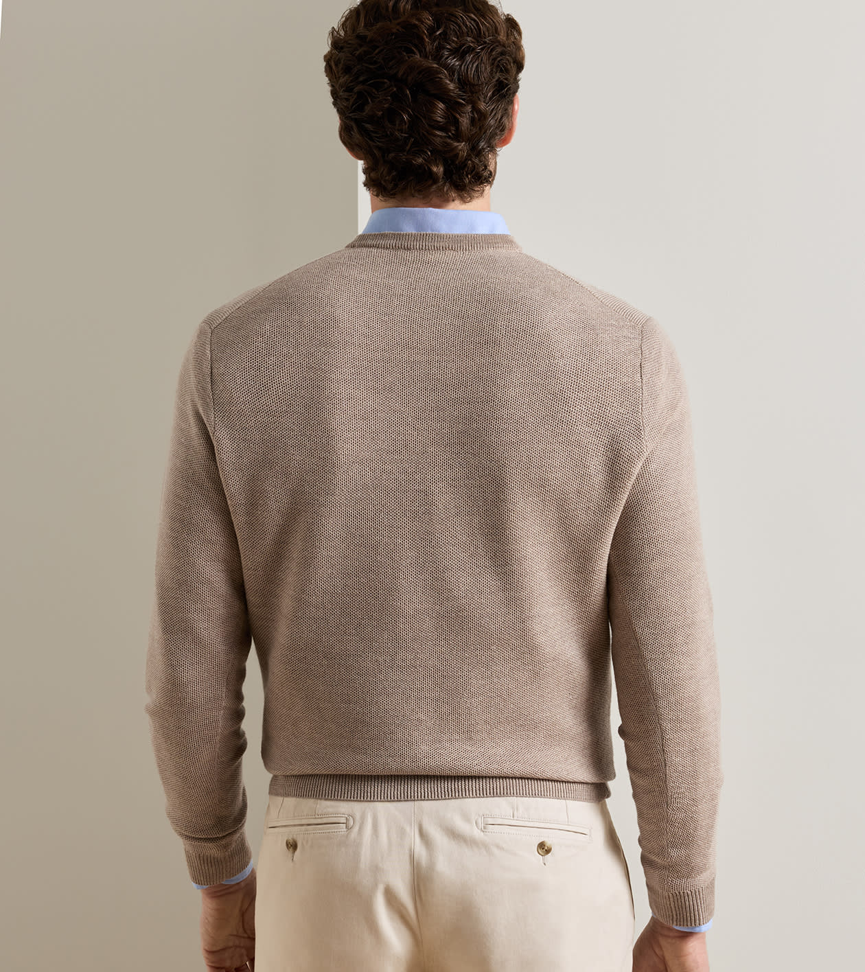 A man viewed from behind wearing a light brown knit sweater over a light blue collared shirt and cream trousers, set against a plain light gray background.