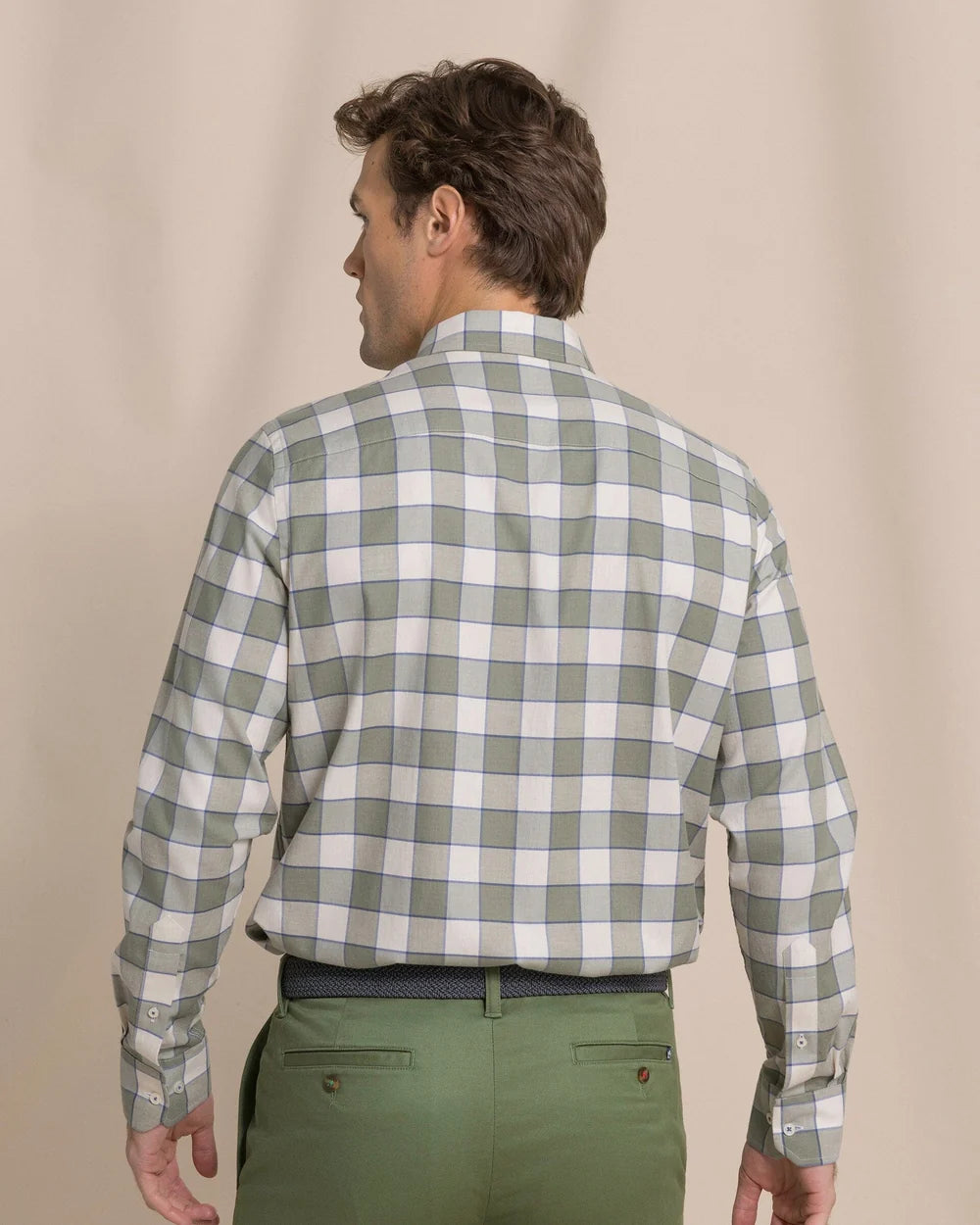 A man viewed from the back wearing a long-sleeve button-up shirt with a green and white check pattern, paired with olive green trousers, against a plain beige studio background.