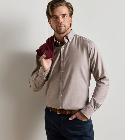 A man wearing a light taupe long-sleeve button-down dress shirt, dark navy trousers, and a brown belt, holding a dark burgundy shirt under his arm against a light gray studio background.