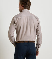 A man viewed from the back wearing a light beige long-sleeve button-down shirt with a subtle vertical pleat at the center back, paired with navy blue trousers and a brown belt, against a plain off-white background.