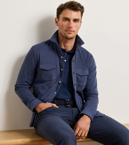 A man wearing a navy blue button-up overshirt, matching navy trousers, and a navy polo shirt, sitting on a wooden bench against a plain off-white wall.