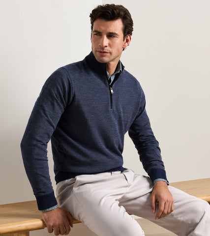 A man wearing a navy blue quarter-zip knit sweater over a light blue collared shirt and off-white trousers, sitting on a wooden bench against a plain light background.