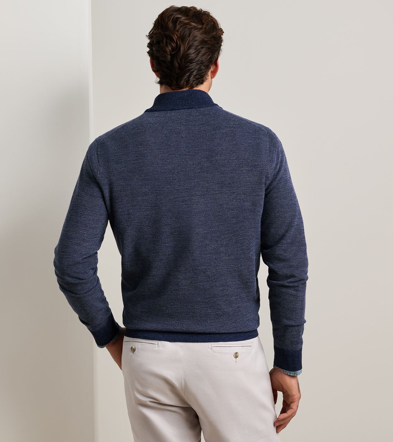 A man viewed from behind wearing a navy blue knit sweater with a ribbed collar, hem, and cuffs, paired with off-white trousers, against a neutral light gray background.