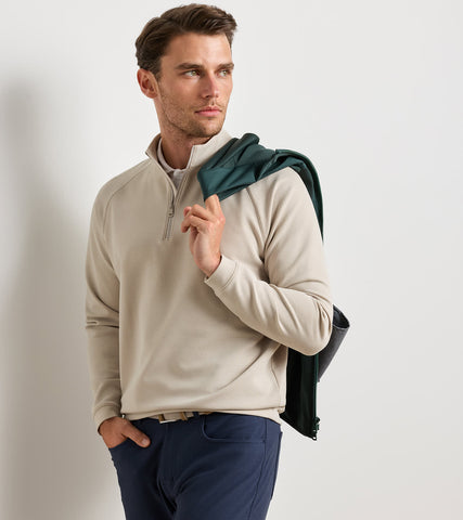 A man wearing a light beige quarter-zip pullover sweater and navy trousers, with a dark green jacket draped over his shoulder, set against a plain light gray studio background.