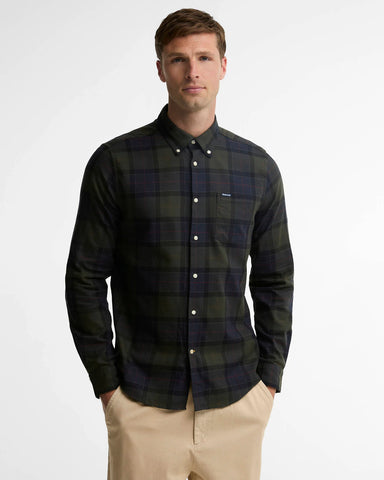 A man wearing a dark green and navy plaid button-down shirt with beige pants, standing against a light gray studio background.