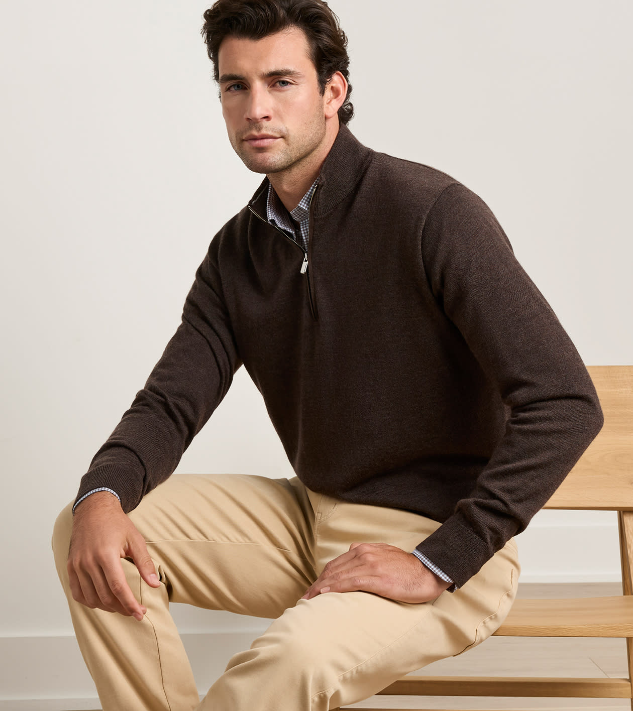 A man wearing a dark brown quarter-zip sweater over a collared shirt and khaki pants, sitting on a wooden bench against a light gray wall.