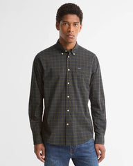 A man wearing a dark olive green and black plaid button-down shirt with a small white logo on the chest, paired with blue jeans, against a light gray background.