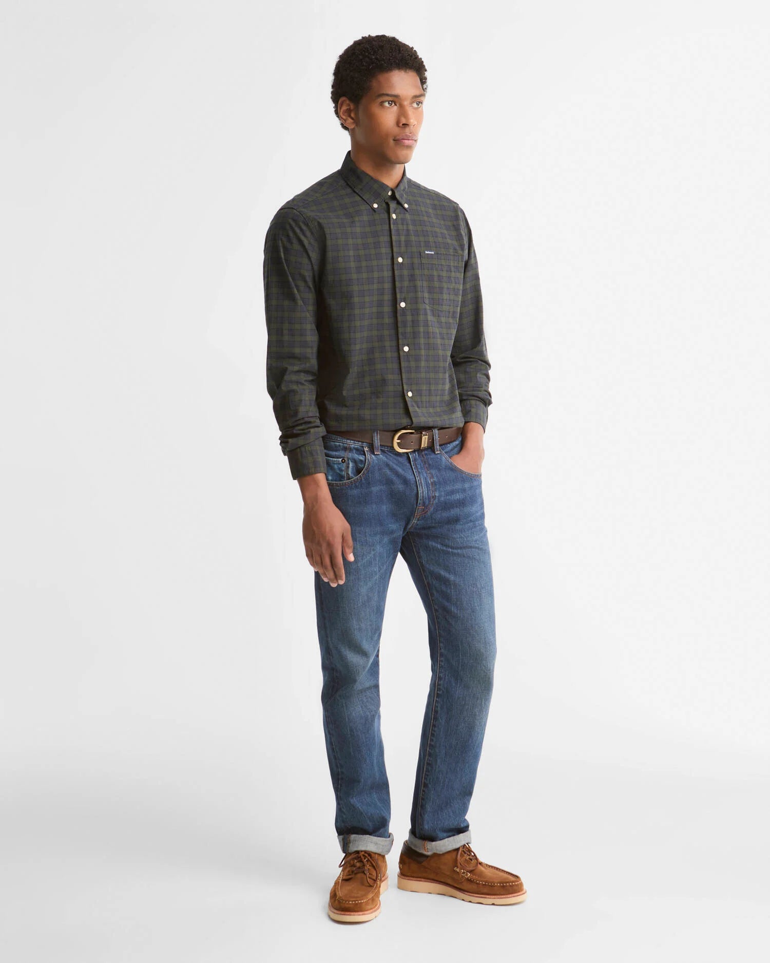 A man modeling a dark gray and green plaid button-down shirt, blue jeans, and brown suede boots against a plain white studio background.