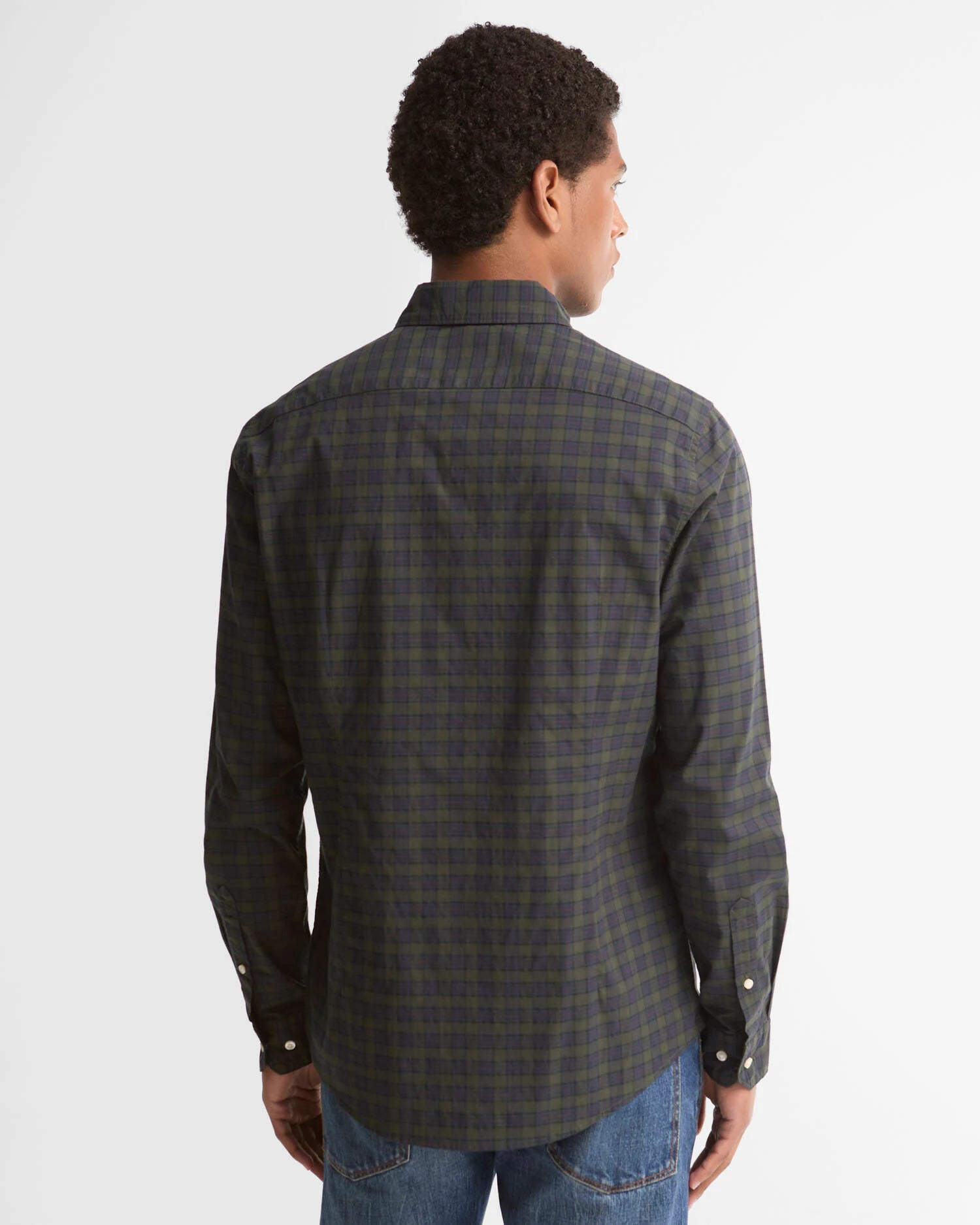 A man viewed from the back wearing a dark green and black plaid button-down shirt with blue jeans, set against a plain light gray studio background.