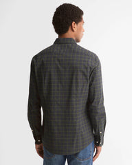 A man viewed from the back wearing a dark green and black plaid button-down shirt with blue jeans, set against a plain light gray studio background.