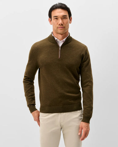 A man wearing a dark olive brown quarter-zip knit sweater over a light pink collared shirt and beige trousers, standing against a light gray studio background.
