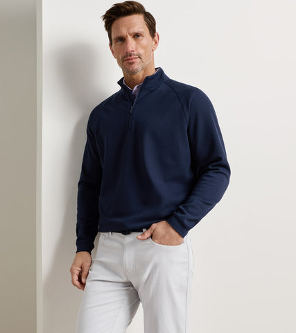 A man wearing a navy blue quarter-zip long-sleeve pullover sweater and light gray trousers, standing against a plain off-white wall.