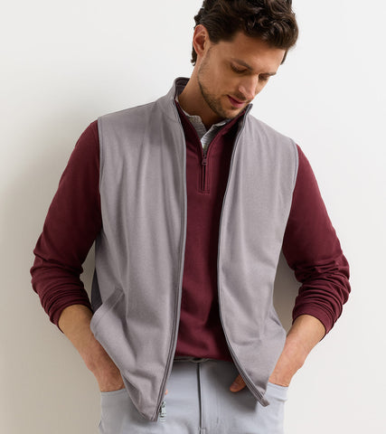 A man wearing a light gray zip-up fleece vest over a burgundy quarter-zip long-sleeve shirt and light gray trousers, standing against a plain off-white wall.